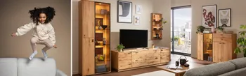 Gemütliches Wohnzimmer mit modernen Holzmöbeln, beleuchteter Vitrine, TV-Lowboard, Wandregal, Sideboard und grauem Sofa, Blick auf Balkon im Hintergrund.
